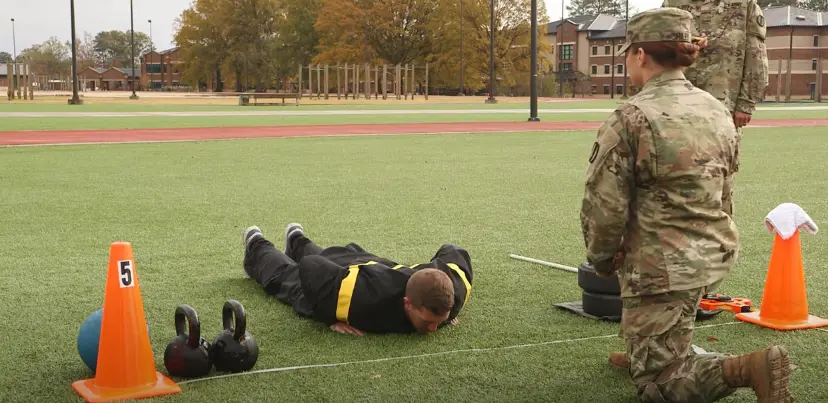 Soldier doing ACFT push-up on grassy field near kettlebells and cone, observed by female military personnel.