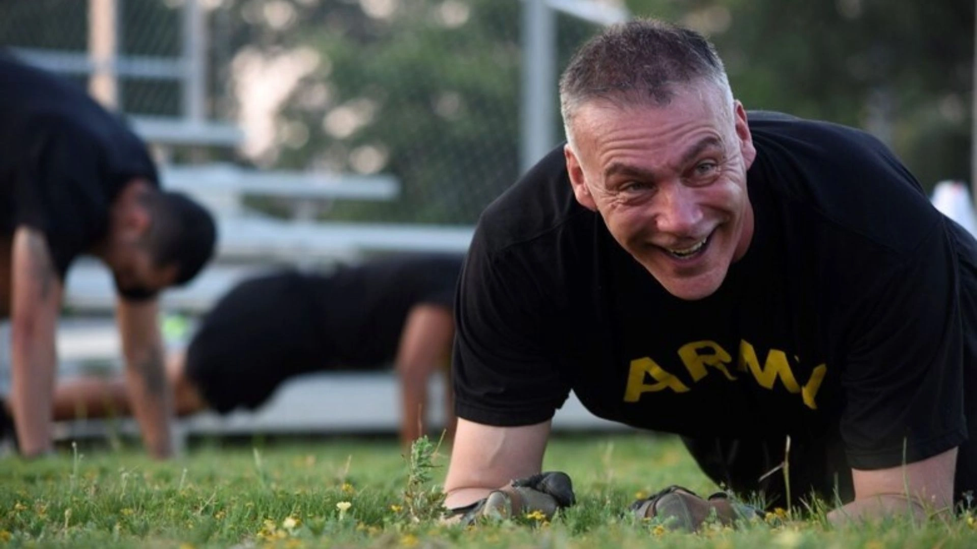ACFT Calculator | Calculate Your Army Combat Fitness Test Score 6 Soldier in a black "ARMY" shirt doing a plank on a grassy field, smiling, with others in the background