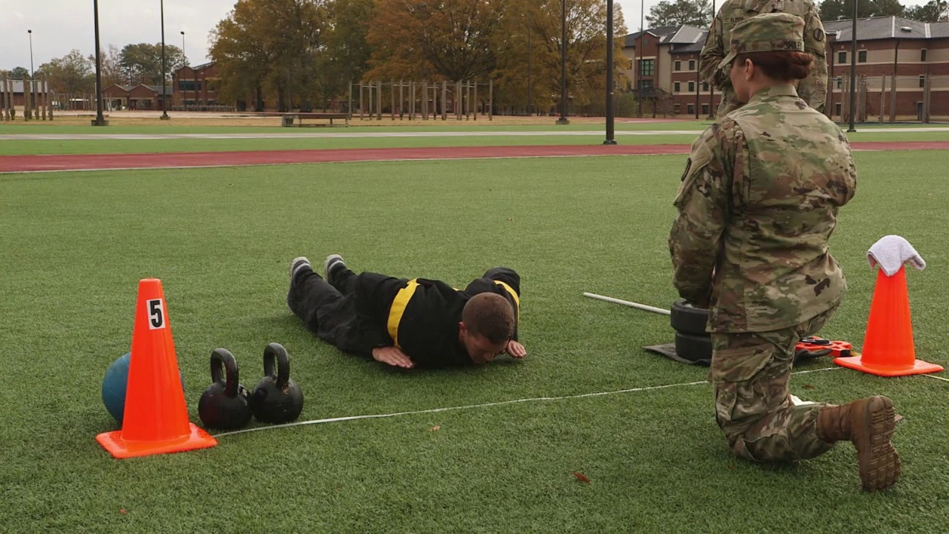 U.S. Army soldier doing push-ups in the HRP event on grass while a female ACFT administrator observes.