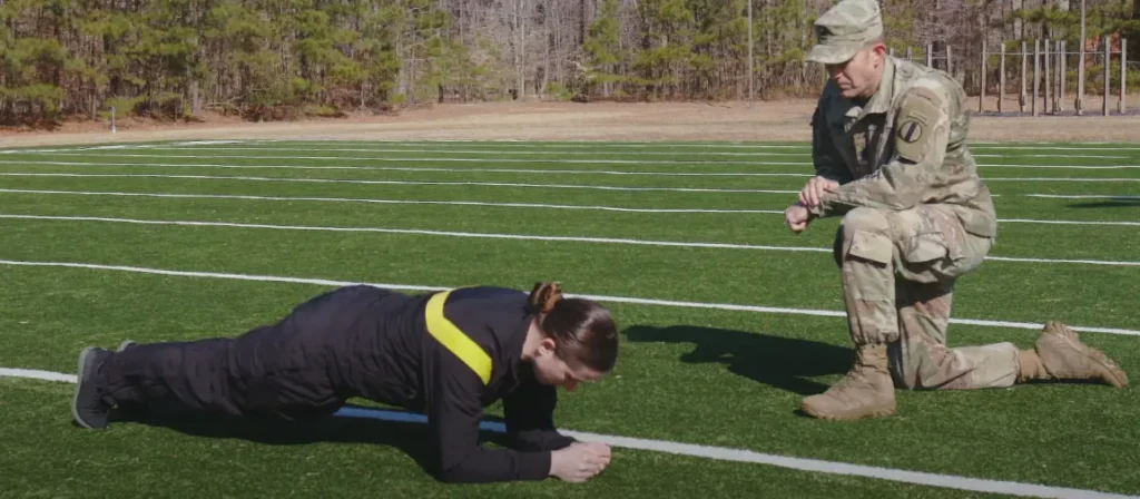 Female soldier doing plank on grassy field observed by military personnel, with trees and obstacle course in background