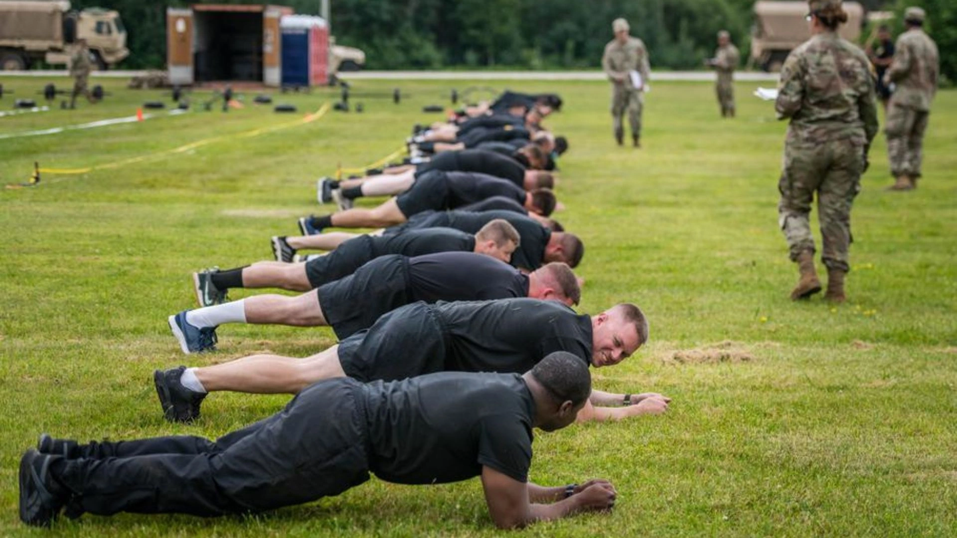 ACFT Calculator | Calculate Your Army Combat Fitness Test Score 4 Soldiers in black attire performing plank (PLK) on a grassy field during ACFT training, with military personnel observing