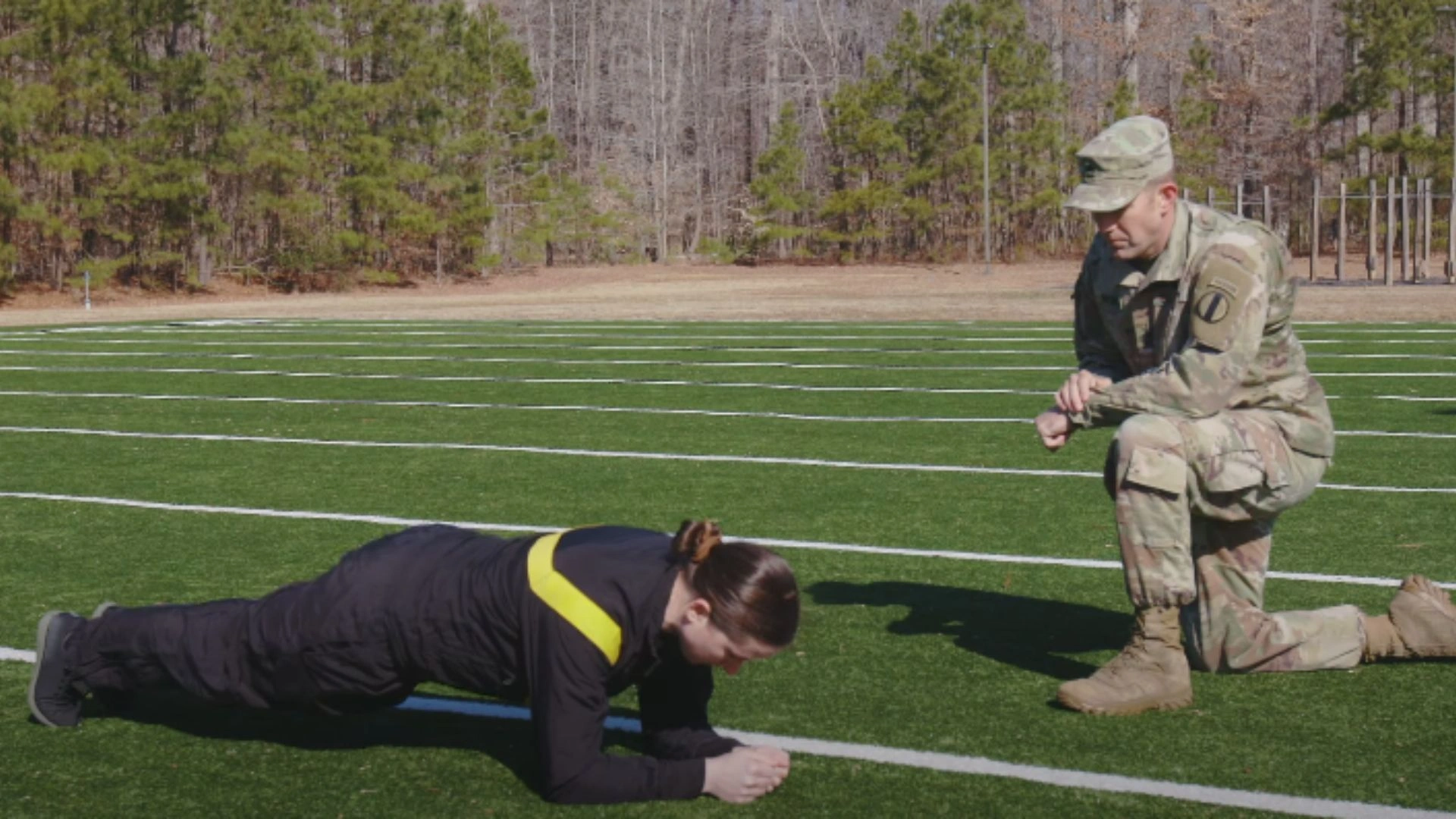 Female U.S. Army soldier planking on grass during the PLK event while an ACFT administrator in uniform kneels nearby.