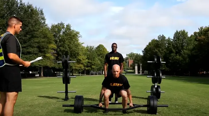 Soldier in black "ARMY" T-shirt performing a trap bar deadlift outdoors, supervised by two others on a grassy field